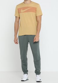 Hombre de pie vestido con una camiseta de Nike mostaza, pantalones deportivos grises y zapatillas Nike blancas frente a un fondo blanco liso.