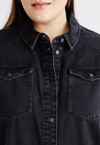 Zwarte denim overhemd met een knoopsluiting, twee borstzakken en contrasterende stiksels. De stof heeft een soepele, gestructureerde uitstraling.