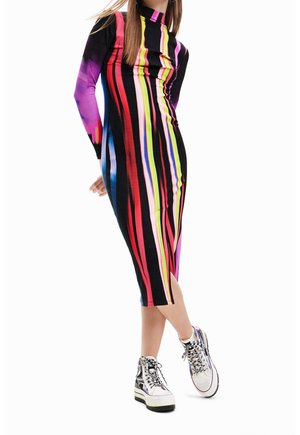 Žena v černých midi šatech s vertikálními vícebarevnými pruhy a bočním rozparkem, doplněná o vysoké tenisky s barevnými tkaničkami.