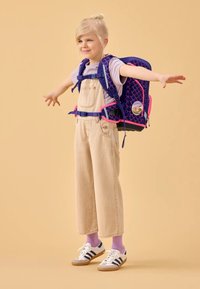 Kind trägt ein lavendelfarbenes T-Shirt und beige Overalls, dazu einen marineblauen und pinkfarbenen Rucksack mit einem Muster aus Meerjungfrauen-Schuppen und einem runden Abzeichen.