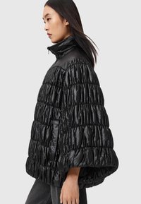 Svart vadderad jacka med hög krage. Har en quilteffekt och en överdimensionerad design. Mjukt, glänsande material med framträdande sömmar.