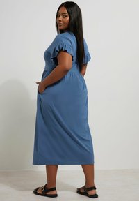 Maxi sukienka z miękkiego niebieskiego materiału, z krótkimi falbaniastymi rękawami, z wcięciem w talii, bocznymi kieszeniami i zwiewnym krojem. Na stopach czarne sandały.