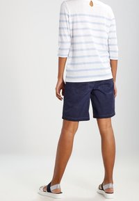 Een persoon die met de rug naar de camera staat, gekleed in een wit en lichtblauw gestreept shirt, marineblauwe knielange shorts en marineblauwe en witte sandalen.