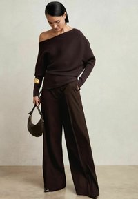 Dunkelbrauner, gerippter Off-Shoulder-Pullover kombiniert mit weiten braunen Hosen. Das Model hält eine kleine, kurvige Handtasche mit goldenen Akzenten.