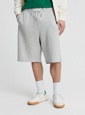 Mężczyzna w jasnoszarych spodenkach z sznurkiem, białych skarpetkach i białych trampkach z zielonymi akcentami, stojący z rękami w kieszeniach.