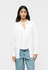 Νεαρή γυναίκα που φοράει ένα λευκό πουκάμισο ανοιχτό πάνω από ένα λευκό μπλουζάκι, συνδυασμένο με ανοιχτό μπλε τζιν, στέκεται μπροστά σε έναν απλό φόντο.