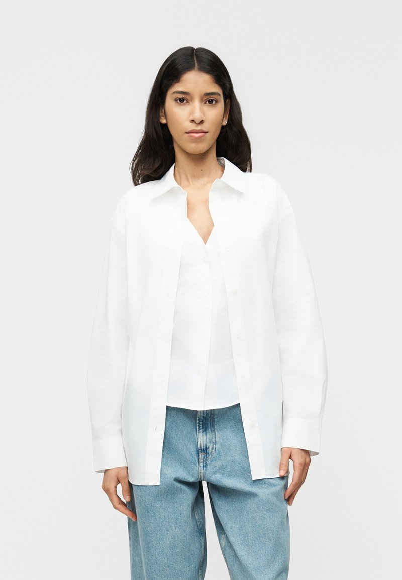 Νεαρή γυναίκα που φοράει ένα λευκό πουκάμισο ανοιχτό πάνω από ένα λευκό μπλουζάκι, συνδυασμένο με ανοιχτό μπλε τζιν, στέκεται μπροστά σε έναν απλό φόντο.