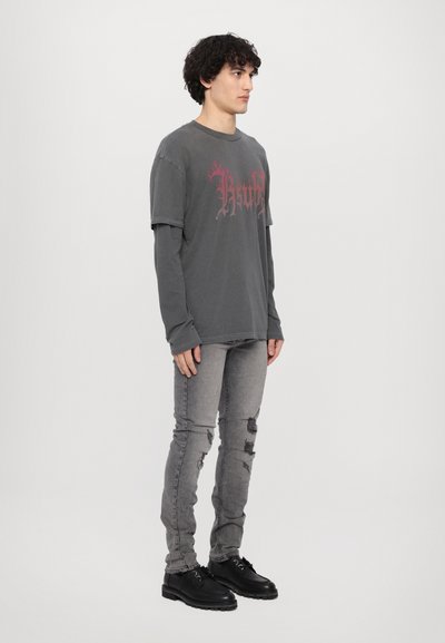 T-shirt oversize gris avec impression graphique rouge superposée sur une manche longue, associée à un jean skinny gris usé et des chaussures à lacets noires.