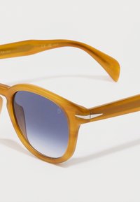 Γυαλιά ηλίου σε χρώμα μελιού με στρογγυλό σκελετό, γαλάζιους φακούς με κλίση και ασημένια λεπτομέρεια στους κροτάφους. Απαλή πλαστική υφή.