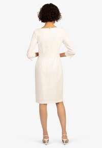 Biała, dopasowana sukienka z dekoltem boat, rękawami 3/4 i zamkiem błyskawicznym z tyłu. Gładka tekstura i długość do kolan.