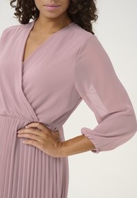 Pleated, light pink dress with a wrap neckline and sheer long sleeves. Features a ruched waist and elasticized sleeve cuffs.