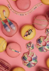 Colorful jewelry arranged on pink macarons: crystal earrings with gradient colors, owl pendant, and a floral design necklace, all sparkling.