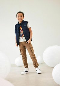 Marinblå pufferväst över en vit grafisk t-shirt, beige cargobyxor med sidofickor och vita sneakers med svarta ränder; minimalisk bakgrund.