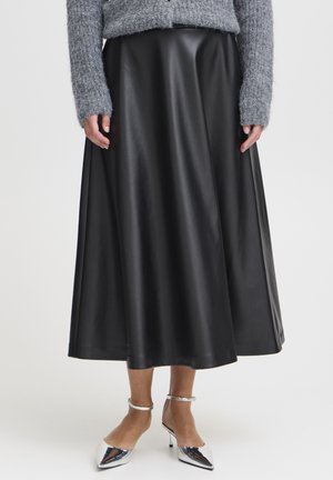 Femme portant une longue jupe en cuir noire, un pull en maille gris et des talons argentés à bout pointu avec bride à la cheville sur fond blanc.