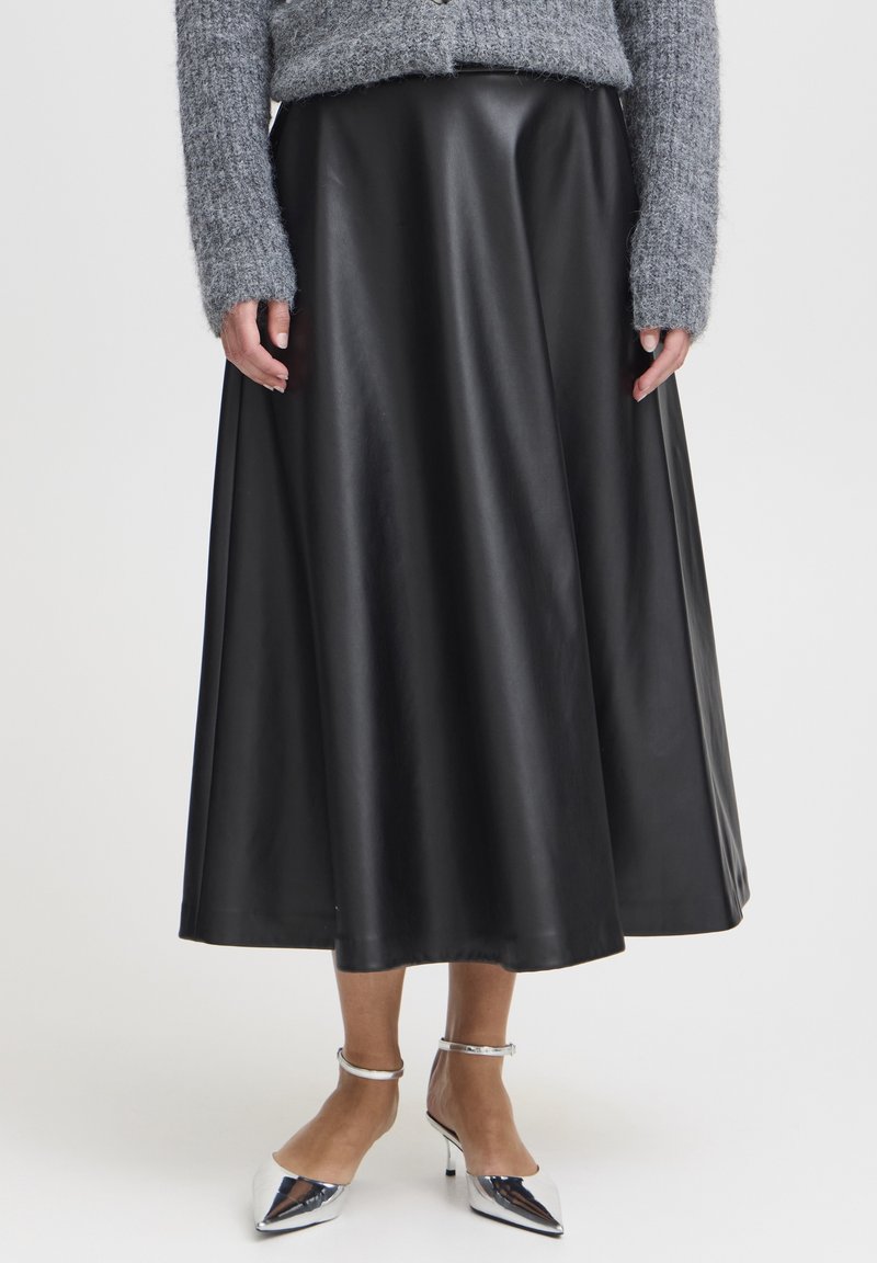 Femme portant une longue jupe en cuir noire, un pull en maille gris et des talons argentés à bout pointu avec bride à la cheville sur fond blanc.