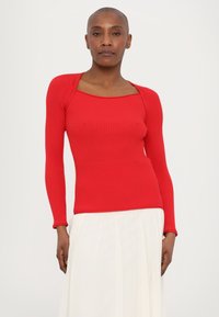 Haut à manches longues rouge à côtes avec un décolleté carré. Design ajusté, texture douce. Associé à une jupe blanche fluide, créant un contraste.