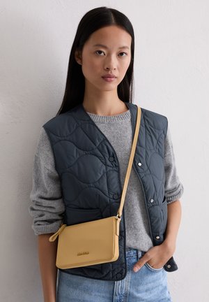 Gequilted marineblauwe vest over een grijze trui, gecombineerd met een kleine gele leren schoudertas. Beschikt over een gladde textuur en een eenvoudig design.