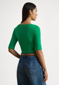 Πράσινο κοντό τοπ με μανίκια μέχρι τον αγκώνα· φορεμένο με ψηλόμεσο τζιν. Λείο ύφασμα, απλός σχεδιασμός και ορατές ραφές στους ώμους.