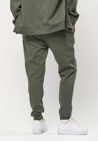 Persona che indossa pantaloni jogger verde oliva e felpa abbinata, abbinati a sneaker bianche, in piedi davanti a uno sfondo uniforme.