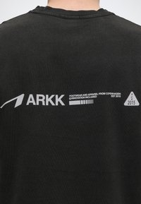 Πλάτη μαύρου πουκαμίσου με λευκό λογότυπο ARKK, κείμενο "Footwear and apparel from Copenhagen, est 2015," και σύμβολο τριγώνου με "AC 2015."