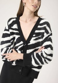 Cardigan cu dungi de zebra alb-negru, cu decolteu adânc în V, mâneci lungi și închidere cu nasturi. Fabricat dintr-un material moale și texturat.