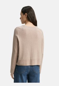 Frau mit langen dunklen Haaren, die einen beigen Pullover und Bluejeans trägt, von hinten vor einem schlichten Hintergrund gezeigt.