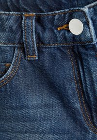 Mörkblå denimjeans med orange sömnad, med en silverknapp och en bältesölj vid midjan. Strukturerad tyg synlig.