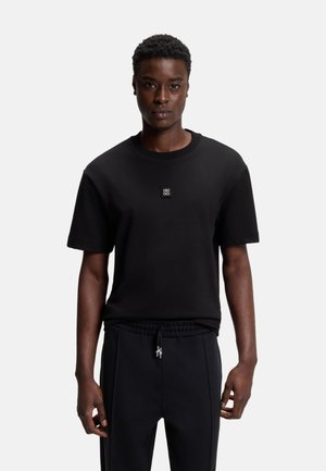 Hombre con camiseta negra de manga corta con pequeño logo "HUGO" en el pecho y pantalones negros con cordón, de pie frente a un fondo claro y liso.