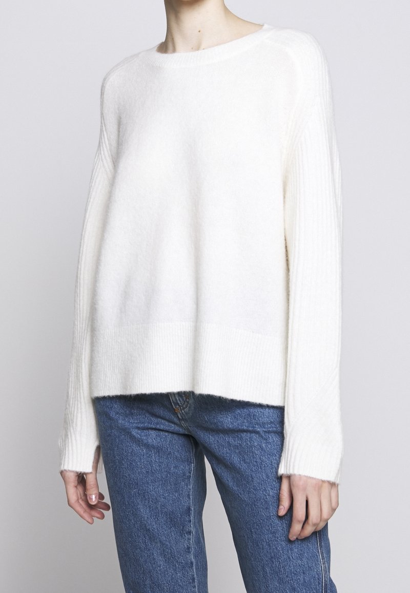 Kremowy sweter z dzianiny prążkowanej o pudełkowym kroju, z okrągłym dekoltem i długimi rękawami. W zestawie z niebieskimi dżinsami.