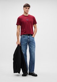 Punane puuvillane t-särk logoga, sinised teksad, mustad nahast kingad ja must jakk, mida hoitakse ühes käes. Ükslane hall taust.