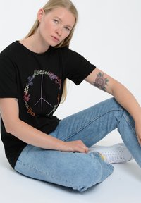 Czarny bawełniany t-shirt z kolorowym wzorem znaku pokoju w postaci kwiatów, w połączeniu z jasnoniebieskimi dżinsami i białymi trampkami, na jednolitym tle.