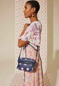 Jūras spēku crossbody soma ar rozā punktiņiem, ar rāvējslēdzēju un regulējamu siksnu. Modelis valkā ziedu raksta kleitu pret rakstainu fonu.