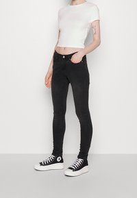 Svarta smala jeans med hög midja och fem fickor, i kombination med en vit kortärmad t-shirt och svarta hög-top sneakers. Slät tygertextur.