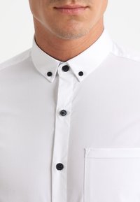 Camicia bianca con colletto classico, bottoni neri e una tasca sul petto. Tessuto liscio con un aspetto pulito e professionale.