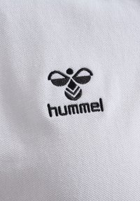 Witte textiel met een zwart geborduurd logo met een bijontwerp boven het woord "hummel." De textuur is glad en geweven.