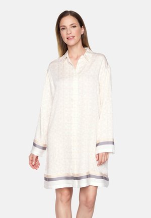 Kobieta ubrana w luźną, kremową sukienkę z długim rękawem, sięgającą kolan, ozdobioną geometrycznymi wzorami oraz szaro-beżowymi paskami na mankietach i dole sukienki.