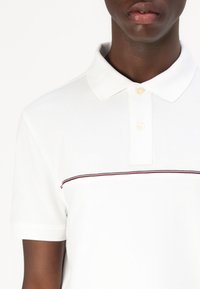 Hvid polo skjorte med et struktureret stof, der har en krave og en knopliste med to knapper. Den har en vandret stribe i rød og blå.