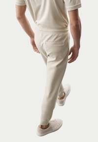 Beige joggerbukser lavet af blødt materiale, med en baglomme, elastisk talje og parret med lyse sneakers.
