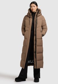 Lang, gevoerd bruin jasje met een hoge kraag en capuchon. Heeft horizontale gestikte secties en een zwarte rits. Draagt over een zwarte outfit.