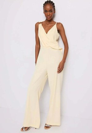 Donna in piedi contro un muro bianco che indossa una tuta senza maniche color crema con pantaloni larghi e tacchi argento open-toe, che guarda avanti con una posa rilassata.