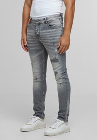 Férfi elhasználódott szürke, kopott skinny farmerben, festékfoltokkal és fehér sportcipőben, egy sima háttér előtt áll.