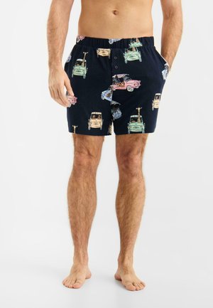 Mandlig torso og ben iført sorte shorts med farverige mønstre af vintagebiler og giraffer, stående barfodet på en hvid baggrund.
