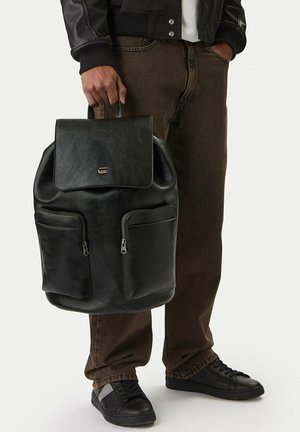 Person trägt braune Hosen und schwarze Schuhe und hält einen schwarzen Leder-Rucksack mit vorderen Reißverschlusstaschen und Klappenverschluss.