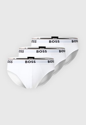 Atletisk mand stående barfodet iført hvide trusser med sort "BOSS" logo på linningen mod ensfarvet hvid baggrund.