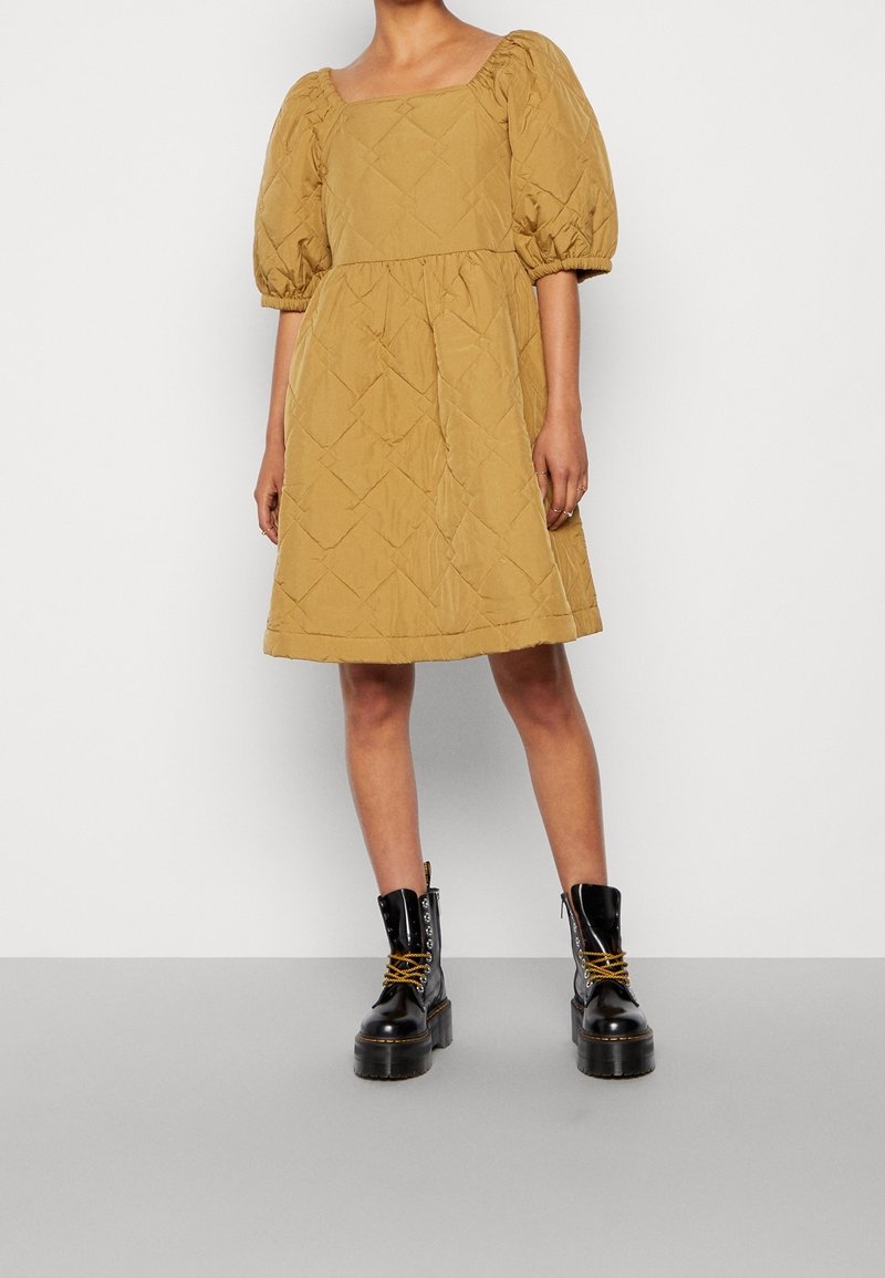 Femme portant une robe matelassée mi-longue couleur moutarde avec des manches bouffantes et des bottines noires à lacets à plateforme, debout sur un sol gris.
