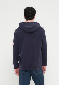 Tmavě modrá mikina s kapucí, klokaní kapsou a kontrastní červeno-bílou vlajkovou nášivkou na levém rukávu. Měkká textura a volný střih.