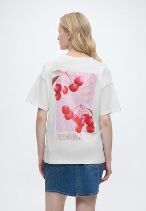 Ξανθιά γυναίκα με λευκό μπλουζάκι με κόκκινο γραφικό κερασάκι και κείμενο "Fresh Harvest", σε συνδυασμό με μπλε τζιν φούστα, εμφανίζεται από πίσω.