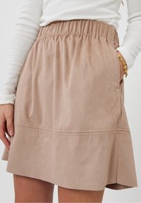 Une jupe en daim beige avec une taille élastique, présente un design évasé et un détail de couture visible à l'ourlet, comporte des poches latérales.
