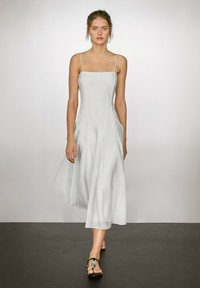 Weißes, ärmelloses Kleid mit geradem Ausschnitt, fließendem Stoff und subtilen Nahtdetails. Getragen mit schwarzen Sandalen mit Riemen.