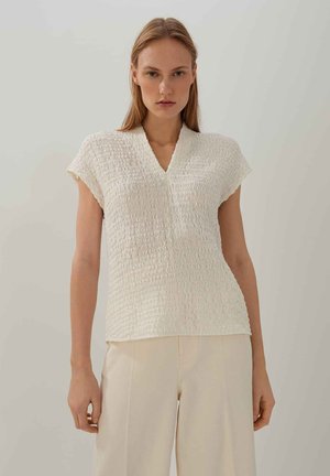 Top bianco a maniche corte con texture, scollo a V e design di tessuto arricciato, abbinato a pantaloni di colore chiaro.